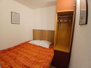 A single bed with a plaid bedspread, a wooden headboard, a white pillow, a wall-mounted clock, and a wooden wardrobe with a mirror. - 2-room apartment - 5 guests, south balcony, L'Alpe d'Huez (Huez)