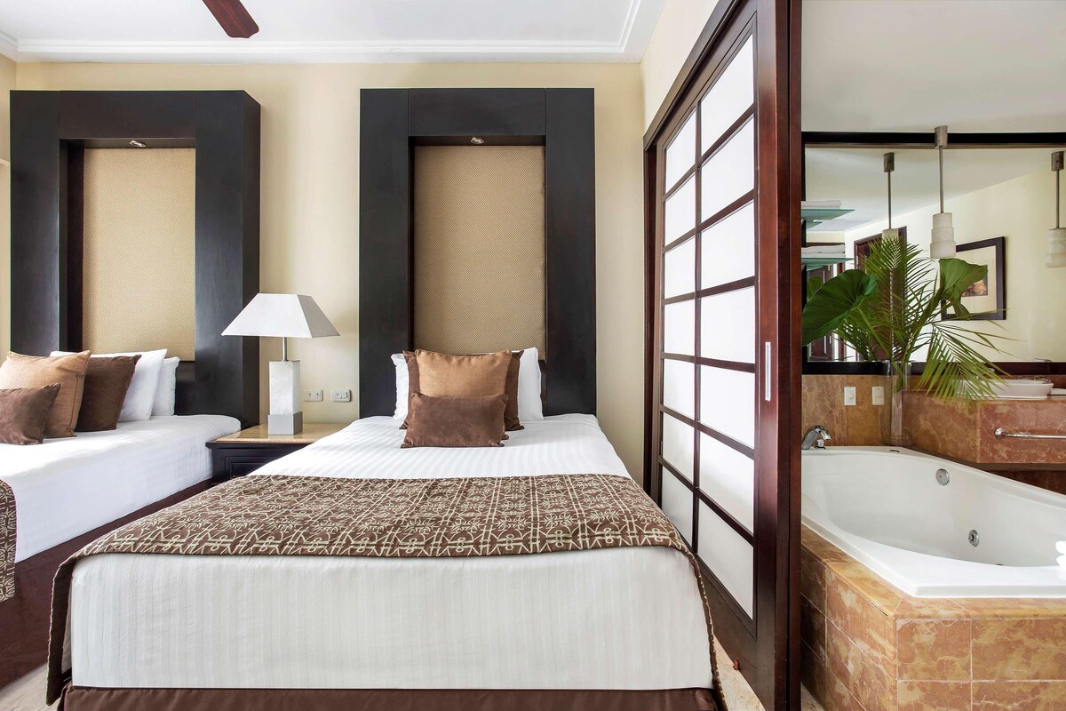 A hotel room with two beds, a large window, a bathroom with a bathtub, and a plant.