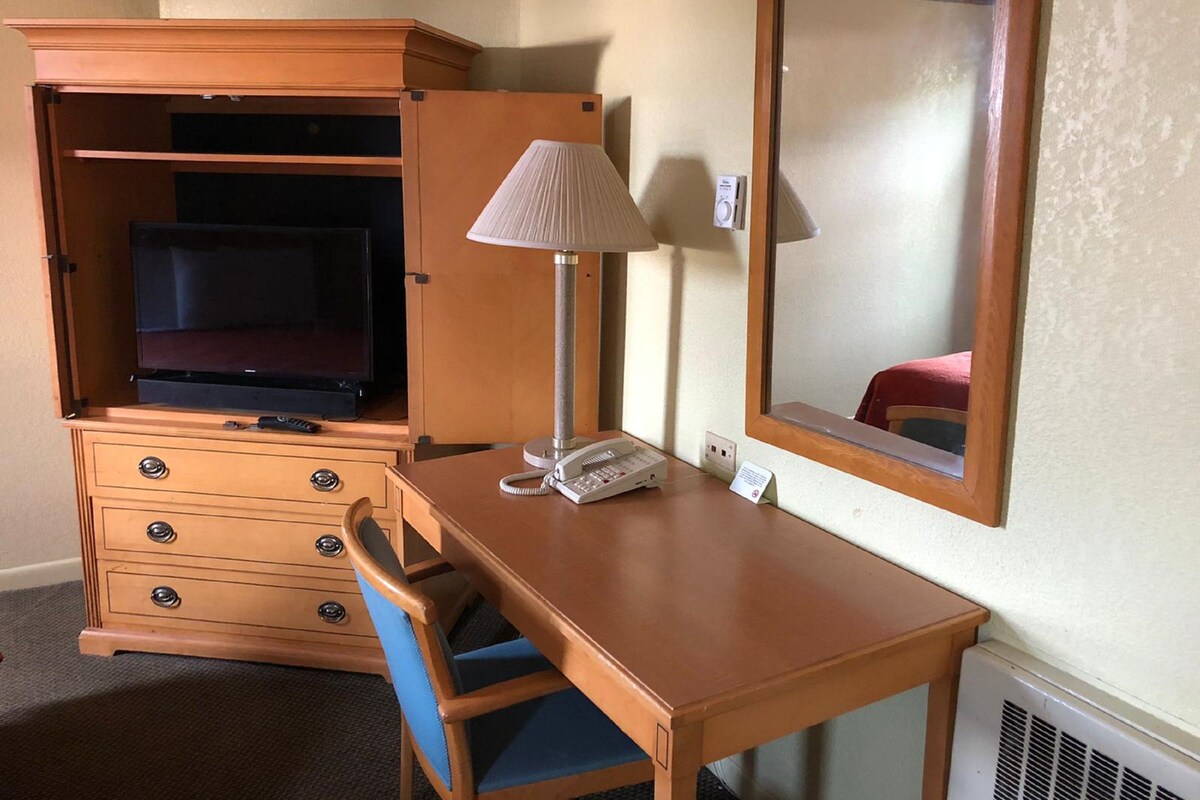 A hotel room with a wooden desk, a telephone, a lamp, a mirror, a TV in a cabinet, and a chair.