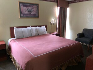 Ein Hotelzimmer mit einem großen Bett, einem Sessel, einem Nachttisch, einer Lampe und einem gerahmten Bild an der Wand.
