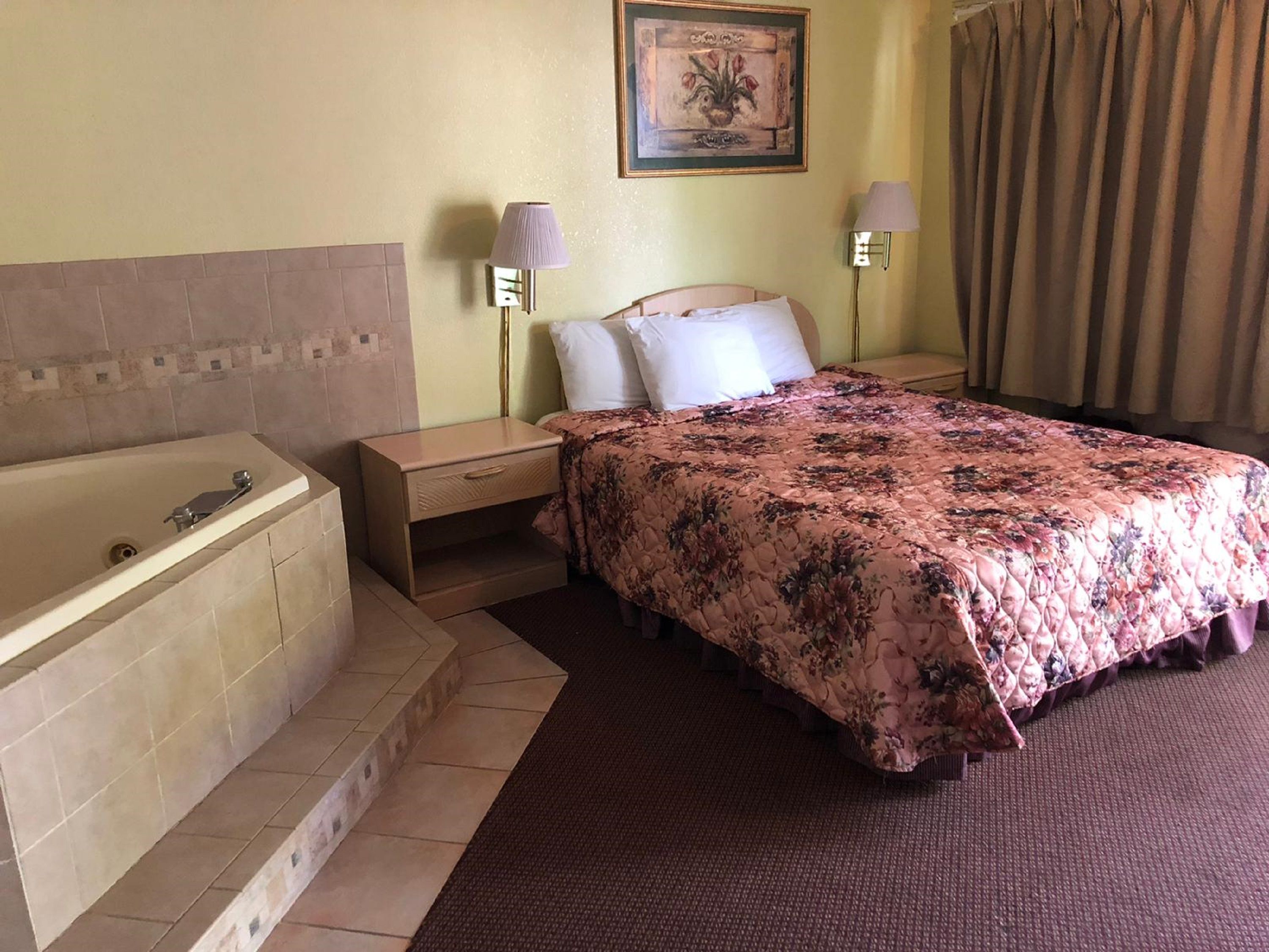 Una camera d'albergo con un letto, comodini, una lampada, un quadro, una tenda e una vasca da bagno.