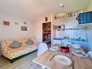 Ein kompakter Wohnraum mit Küchenzeile, einem Esstisch für zwei Personen, einer Couch mit Kissen und einem Hochbett im Hintergrund.