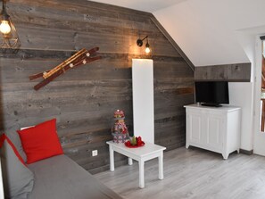 Ein gemütliches Wohnzimmer mit schrägverdeckter Decke, einem Fernseher auf einem weißen Schrank, einem kleinen Tisch mit einem dekorativen Gegenstand und einem roten Kissen auf einem grauen Sofa.