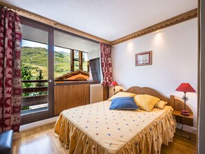 A bedroom with a large bed, a window with curtains, a wooden headboard, and a view of a mountainous landscape. - Bright apartment at the foot of the slopes - 4 guests (Les Menuires)