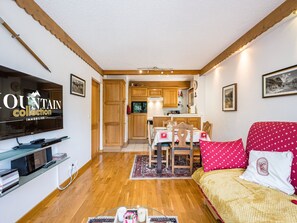 A furnished living room with a flat-screen TV, wooden flooring, a dining area with a table and chairs, and a kitchen in the background. - Bright apartment at the foot of the slopes - 4 guests (Les Menuires)
