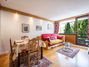 Um quarto aconchegante com uma mesa de jantar posta para dois, um sofá com almofadas vermelhas e vista para uma floresta através de uma grande janela.