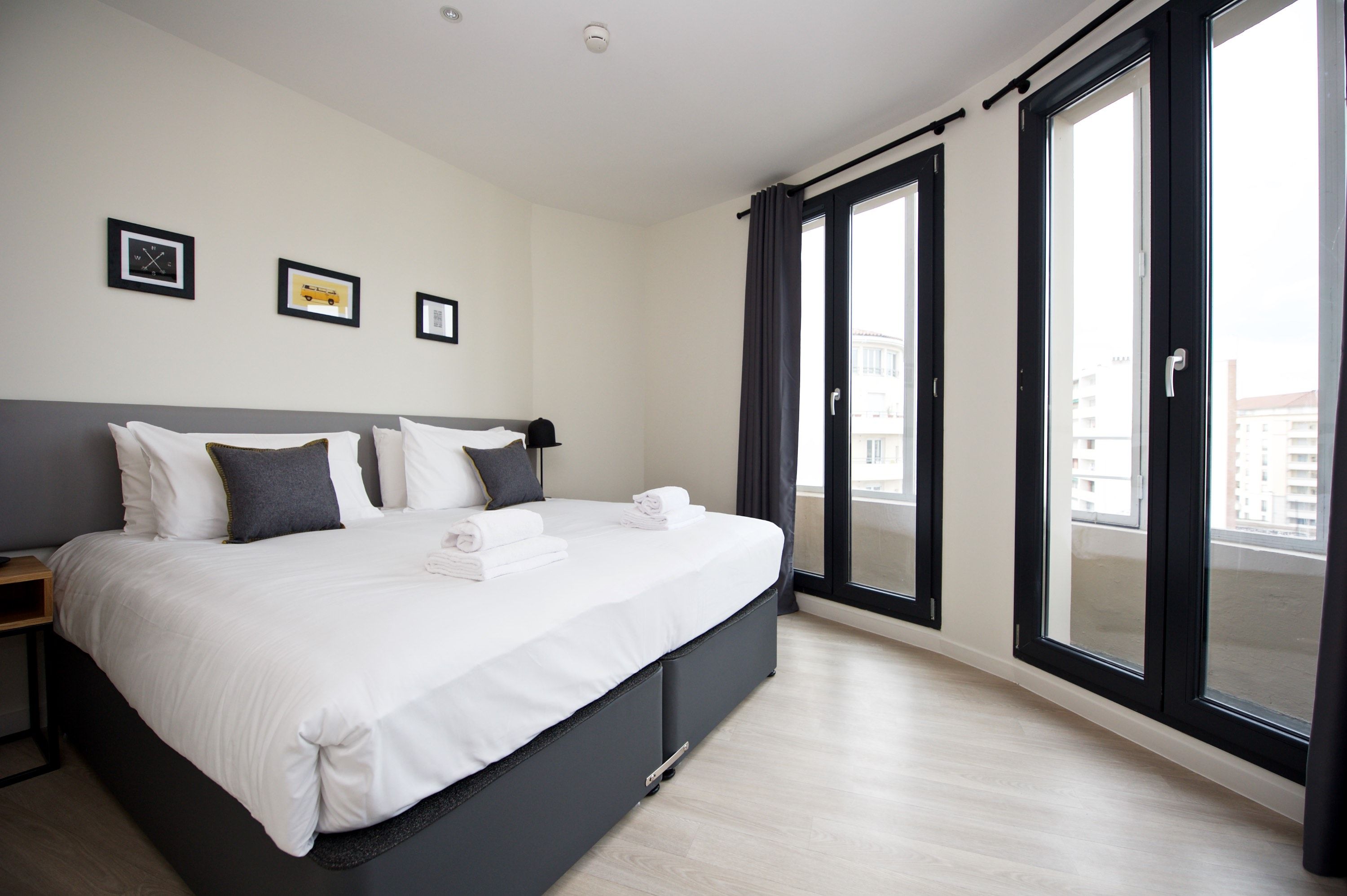 A modern hotel room with a large bed, a bedside table, and a view of the city through floor-to-ceiling windows.