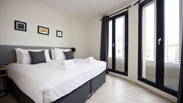 A modern hotel room with a large bed, a bedside table, and a view of the city through floor-to-ceiling windows.