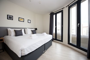 Una habitació d'hotel moderna amb un llit gran, una taula de nit i vistes a la ciutat a través de finestres del sostre al terra.