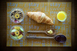 Desayuno con un croissant, ensalada de frutas, granola y un vaso de zumo de naranja sobre una base americana amarilla con la palabra 'stay' repetida.