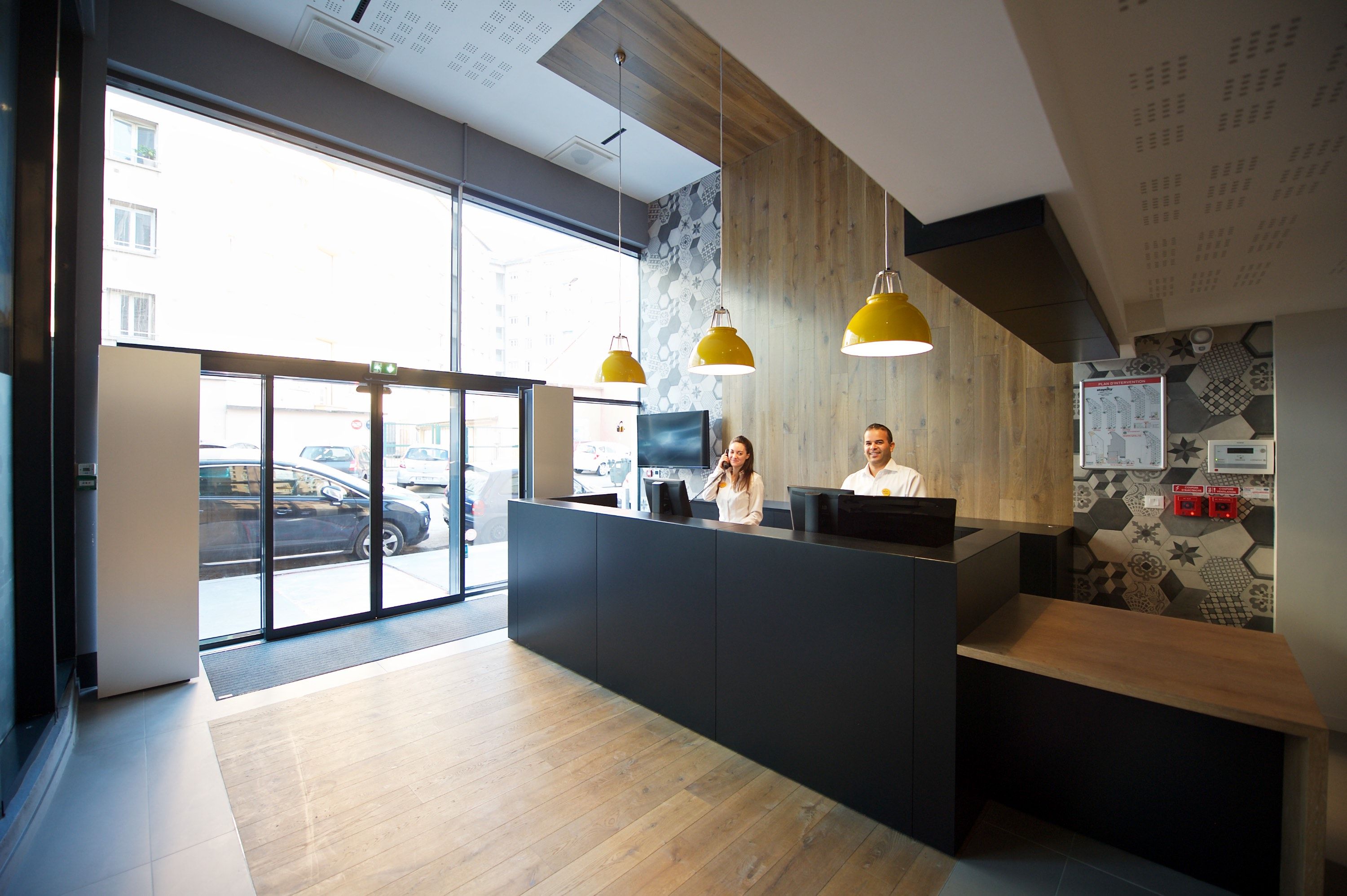 A reception area with a wooden reception desk, two staff members, and a large window overlooking the street.