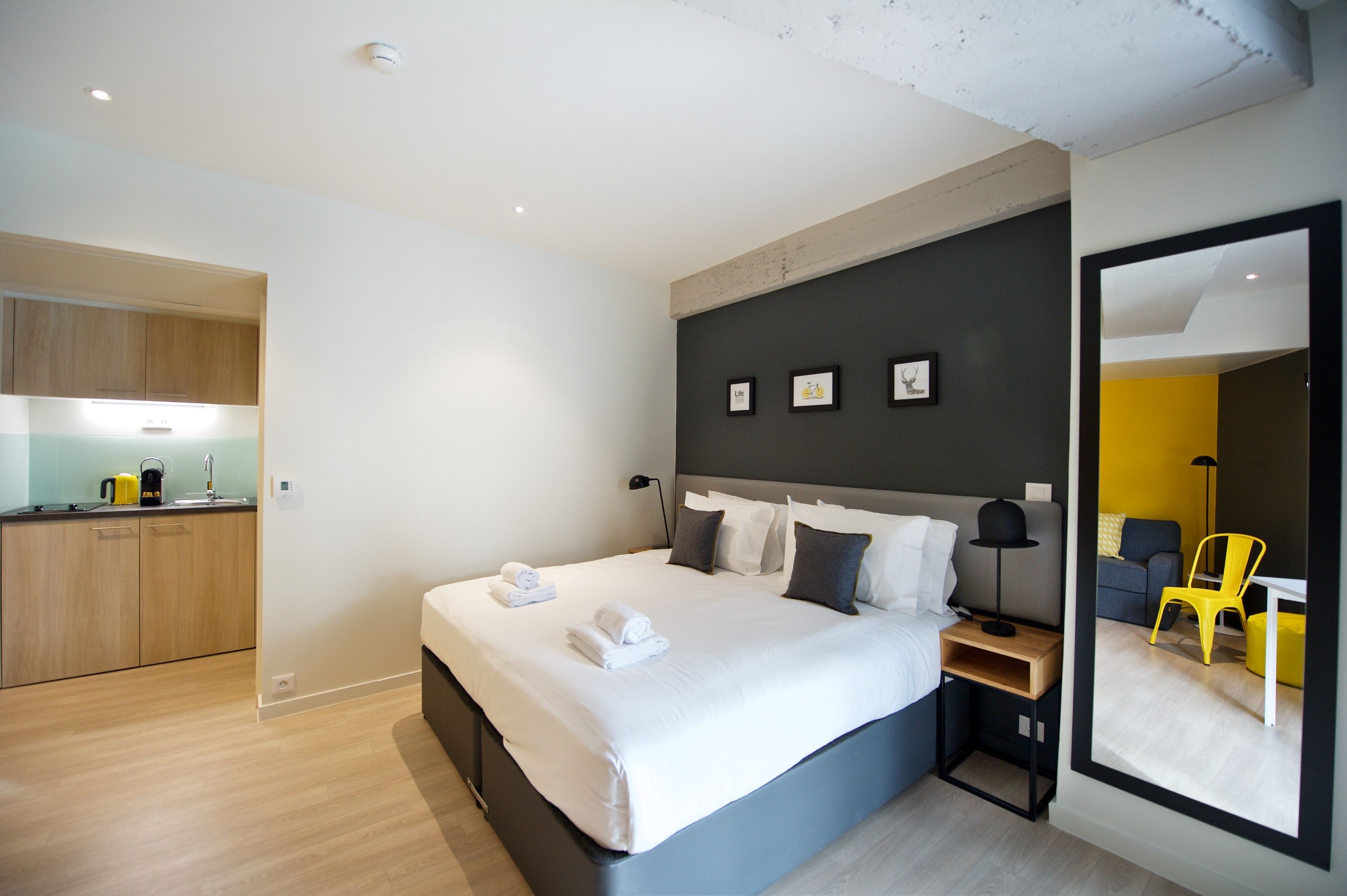 A modern hotel room with a large bed, bedside tables, a mirror, and a view into a dining area.