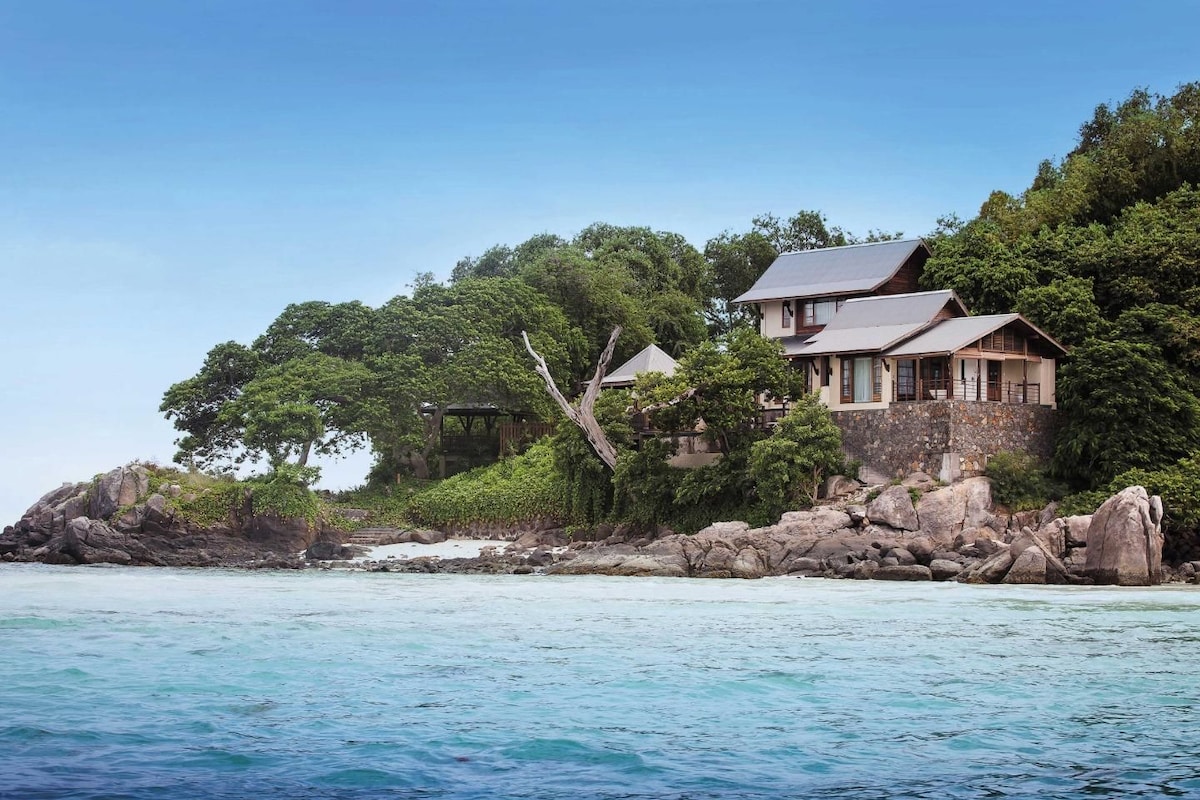A coastal house with a stone foundation, surrounded by lush greenery and overlooking turquoise waters.