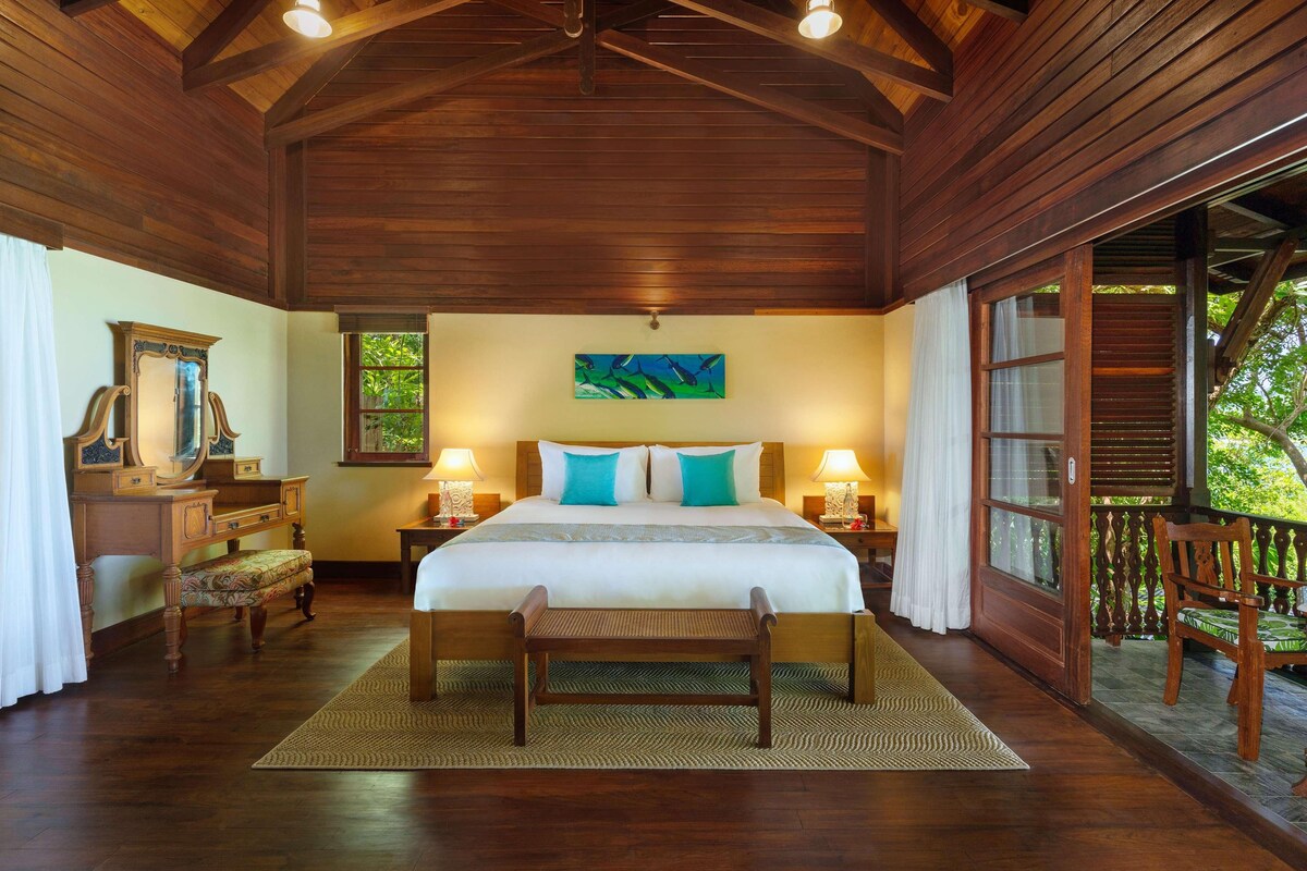 A wooden-paneled bedroom with a large bed, bedside lamps, a bench, and a view of the outdoors.