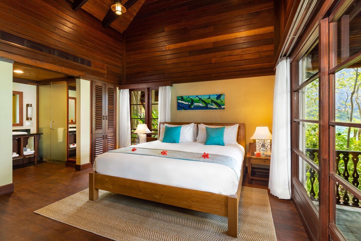 A wooden-framed bedroom with a bed, a painting, and a view of greenery.