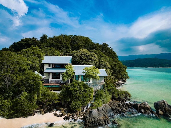 Una casa de dos pisos con balcón, rodeada de exuberante vegetación y vista a un mar turquesa y transparente.