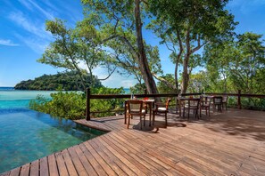 A wooden deck with tables and chairs overlooking a clear blue sea and a tropical island. - JA Enchanted Island Resort (Round Island)