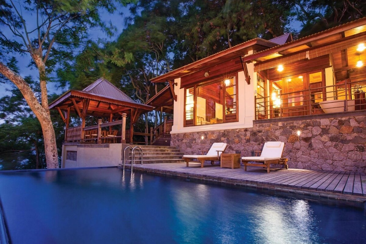 A pool with lounge chairs, a wooden deck, and a stone wall.