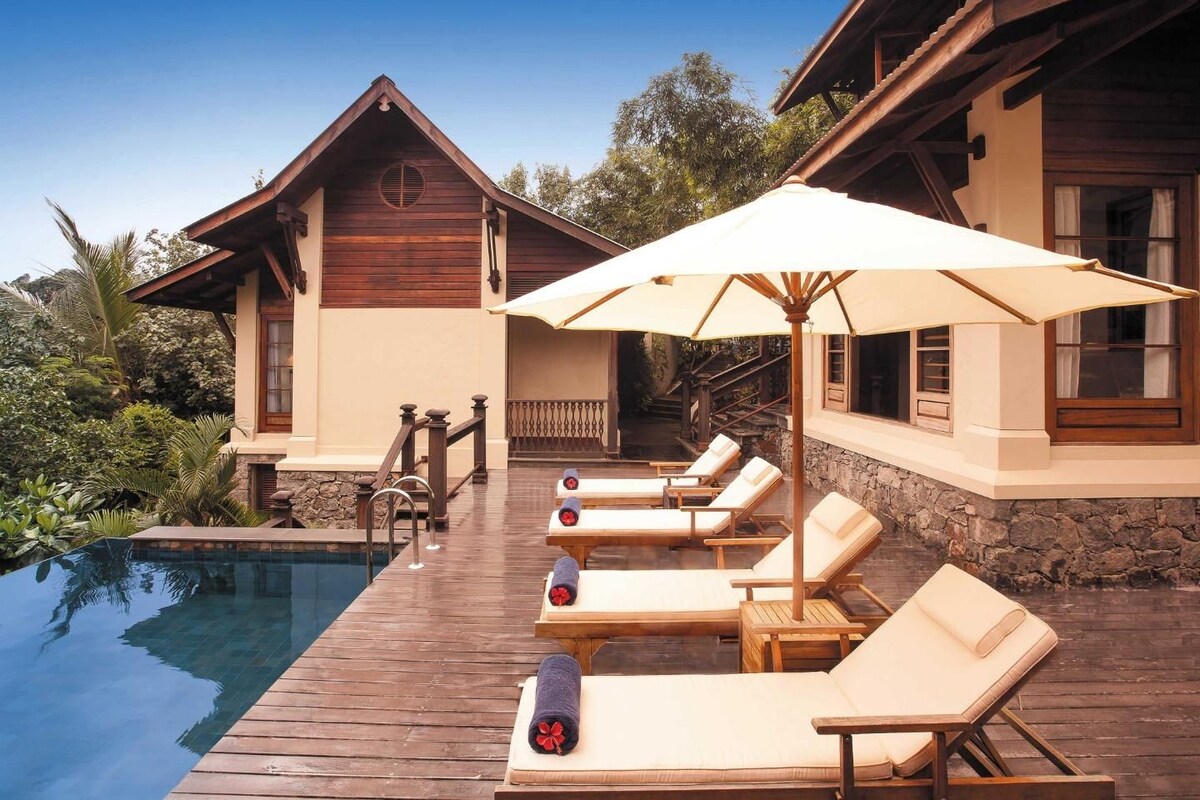 A poolside area with lounge chairs and an umbrella, wooden architecture, and a clear blue sky.