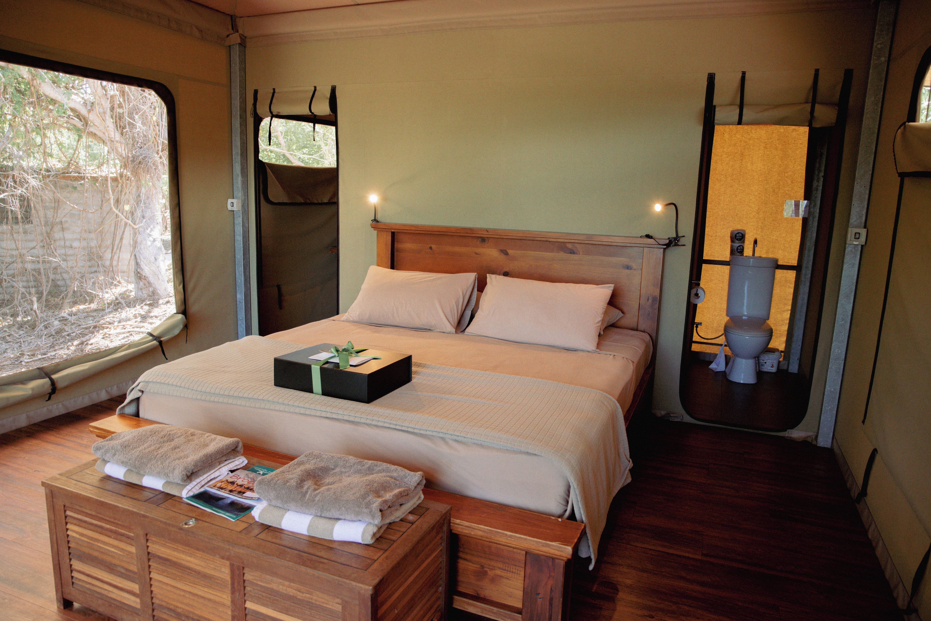 Una cama individual con cabecera de madera, una mesita de noche con un libro y una toalla, y una ventana con vistas a árboles.