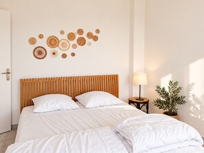 A bedroom with a wooden slatted headboard, white bedding, a wall-mounted circular wall art, a bedside lamp, and a potted plant. - Apartment panoramic view - 6 guests (Chamrousse)