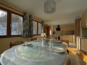 Un coin repas avec une table ronde dressée pour quatre personnes, un vase rempli de fleurs et une vue sur la forêt, visible à travers de grandes fenêtres.