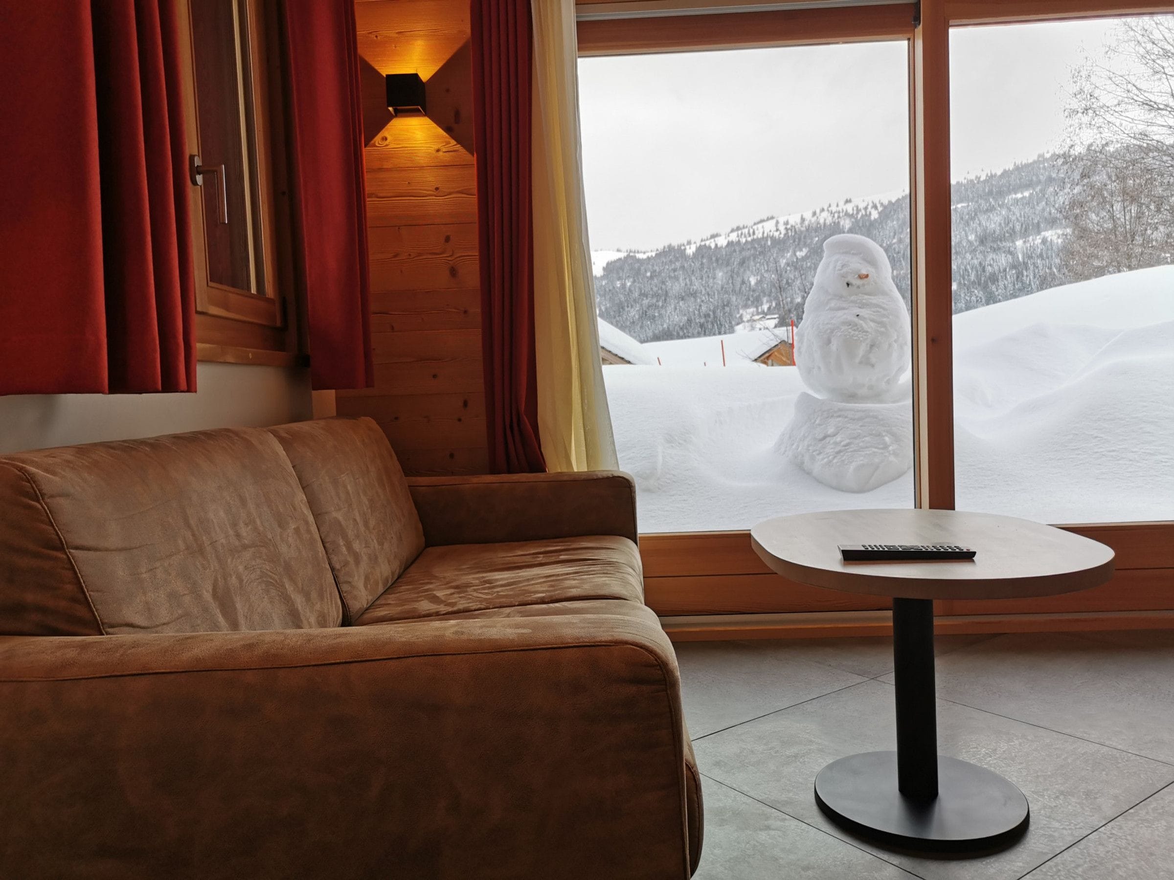 A room with a brown leather sofa, a round table with a remote control, and a large window with a snowy mountain view.