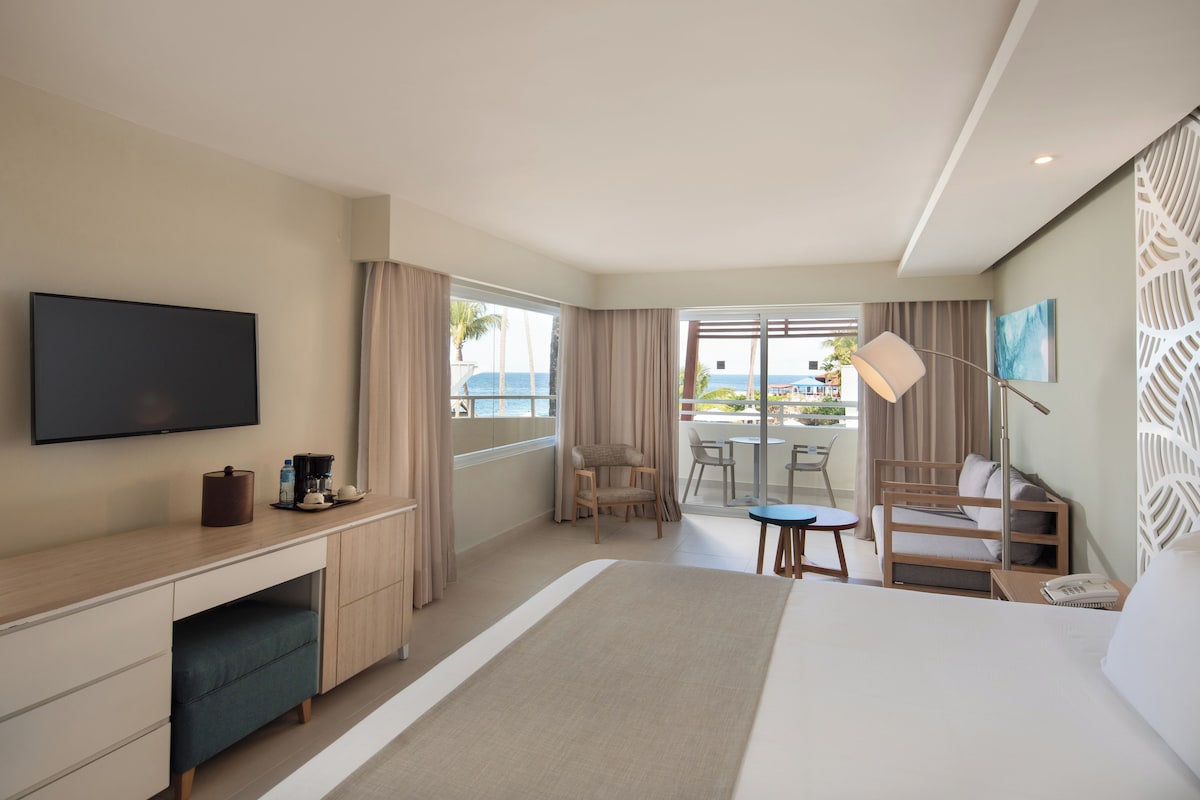 A hotel room with a bed, a desk, a chair, a small table, a TV, and a balcony with a view of the sea.