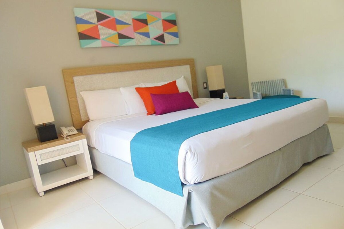 Uma cama bem arrumada com um edredom azul e branco, duas luminárias de cabeceira, uma pequena mesa de cabeceira com um telefone e um quadro vibrante com um padrão geométrico na parede.