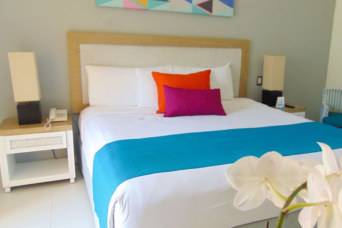 Uma cama bem arrumada com colcha azul e branca, dois travesseiros em laranja e roxo, e uma orquídea branca sobre a cama.