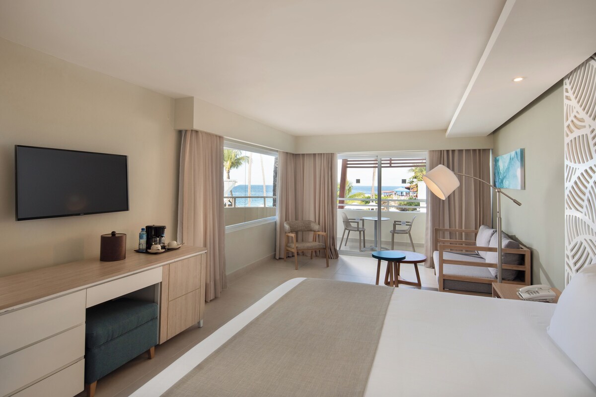 Quarto de hotel com cama, escrivaninha, cadeira, mesinha, televisão e varanda com vista para o mar.