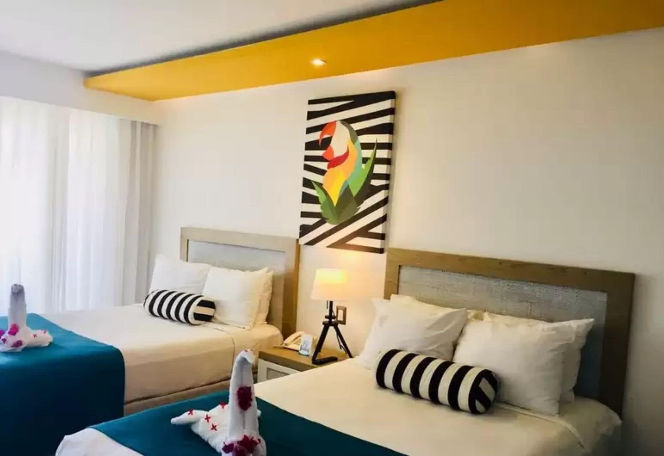 Quarto de hotel com duas camas, cada uma com almofada listrada e manta azul-esverdeada. Há uma obra de arte na parede acima das camas e uma mesa de cabeceira com abajur e telefone.