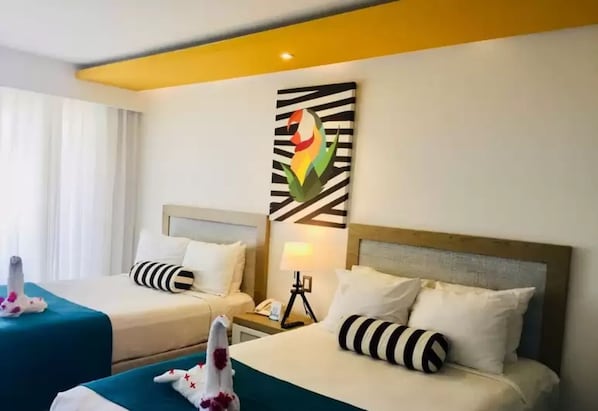 Quarto de hotel com duas camas, cada uma com almofada listrada e manta azul-esverdeada. Há uma obra de arte na parede acima das camas e uma mesa de cabeceira com abajur e telefone.