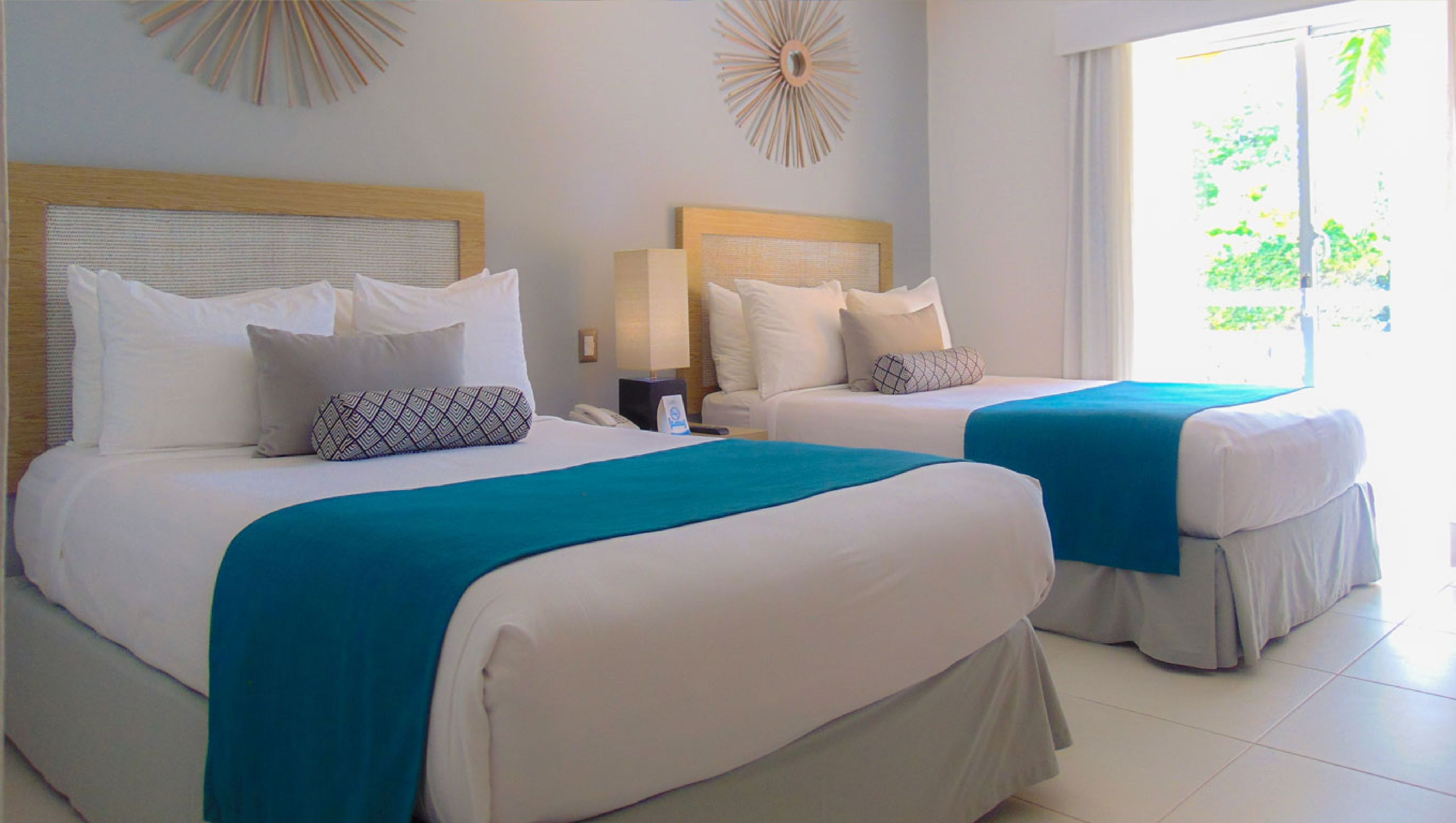 Deux lits jumeaux avec une literie blanche et des plaids turquoise, une tête de lit en bois et une décoration murale en forme de soleil.