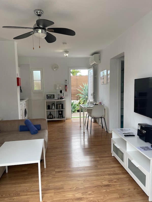 Una sala moderna con ventilador de techo, un comedor con mesa y sillas, un televisor sobre un mueble blanco, y una cocina equipada.