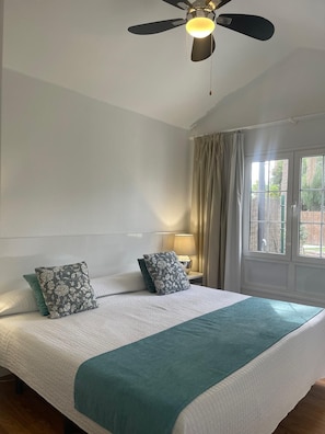 Una habitación de hotel con cama, ventilador de techo, ventana con cortinas y mesita de noche con lámpara.