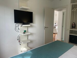 A bedroom with a bed, a television mounted on a white wall, and a door leading to another room.