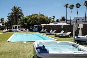 Piscina con sillas de descanso y un jacuzzi, rodeada de palmeras y una cerca de madera.