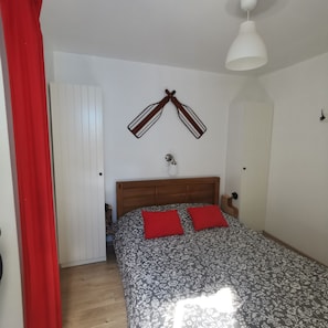 Ein Schlafzimmer mit einem hölzernen Kopfende, einem Bett mit blumigem Bettbezug, zwei roten Kissen, einer weißen Deckenleuchte und einem an der Wand befestigten Handtuchhalter.