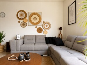 A living room with a grey sectional sofa, a round coffee table with decorative items, and wall-mounted woven art pieces. - Nómada Living | 2 bedrooms, pool, and excellent location (Zaru)