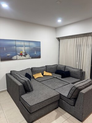 Ein graues Ecksofa mit mehreren Kissen, ein großes Gemälde einer Küstenlandschaft und ein Fenster mit Vorhängen.