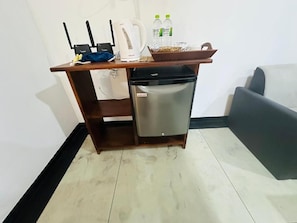 A compact kitchenette with a refrigerator, a kettle, and a coffee maker. There are two bottles of water and a basket on the counter. - Peak Blooms (Ella)