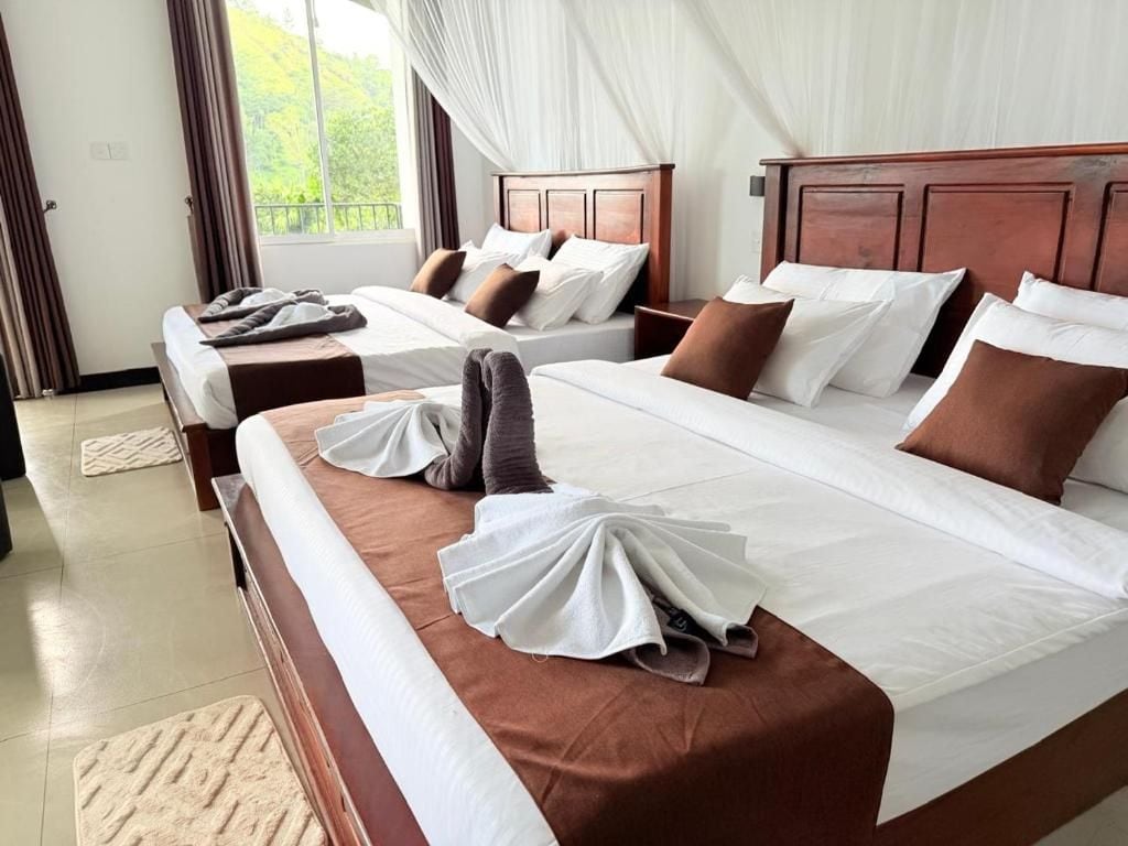 A hotel room with two beds, each with white linens and brown pillows, and a view of greenery through the window.