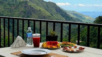 A dining table with plates of food, a water bottle, and a glass of red drink, set against a backdrop of lush green mountains.