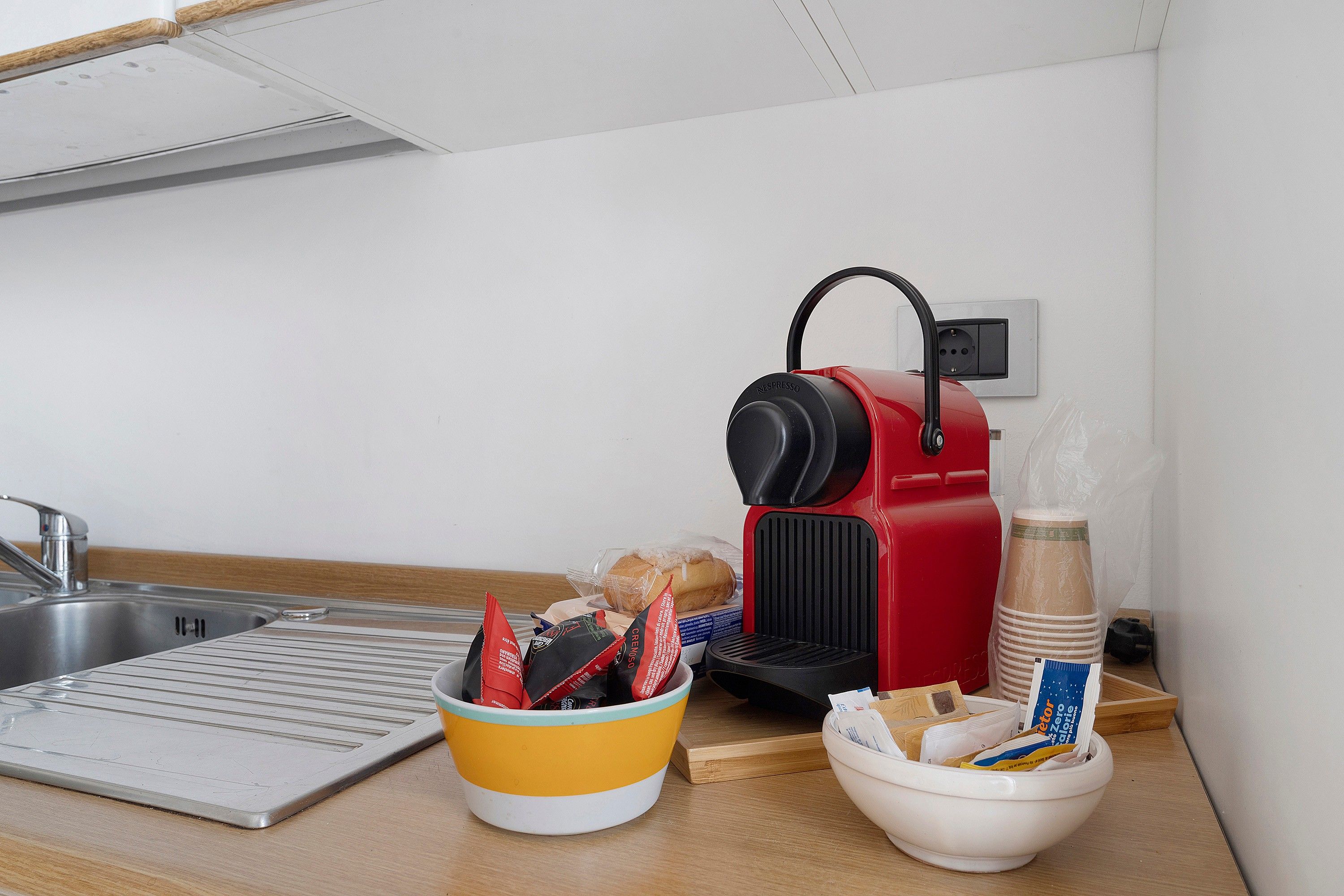 Eine Küche mit einer roten Espressomaschine, einem gelb-weißen Kaffeemühle, einer weißen Schüssel für Kapseln und einem Edelstahl-Spülbecken.