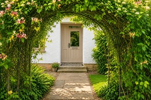A house entrance with a door, surrounded by lush greenery and a stone pathway. - Groom's Cottage, Jedburgh (Jedburgh)