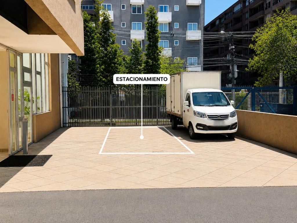 Uma área de estacionamento fechada por grades, com uma van branca estacionada no interior, uma placa indicando 'ESTACIONAMIENTO' e um edifício com várias janelas ao fundo.
