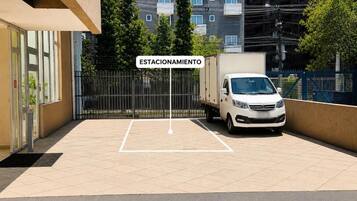 Uma área de estacionamento fechada por grades, com uma van branca estacionada no interior, uma placa indicando 'ESTACIONAMIENTO' e um edifício com várias janelas ao fundo.