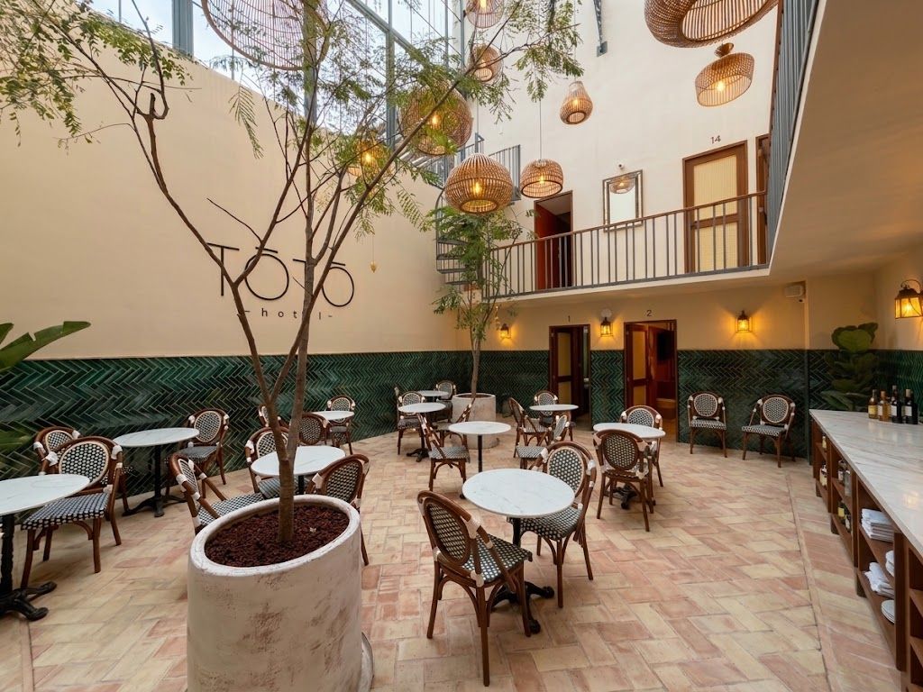 A hotel lobby with multiple seating areas, including round tables and chairs, a large potted tree, and hanging wicker lamps.