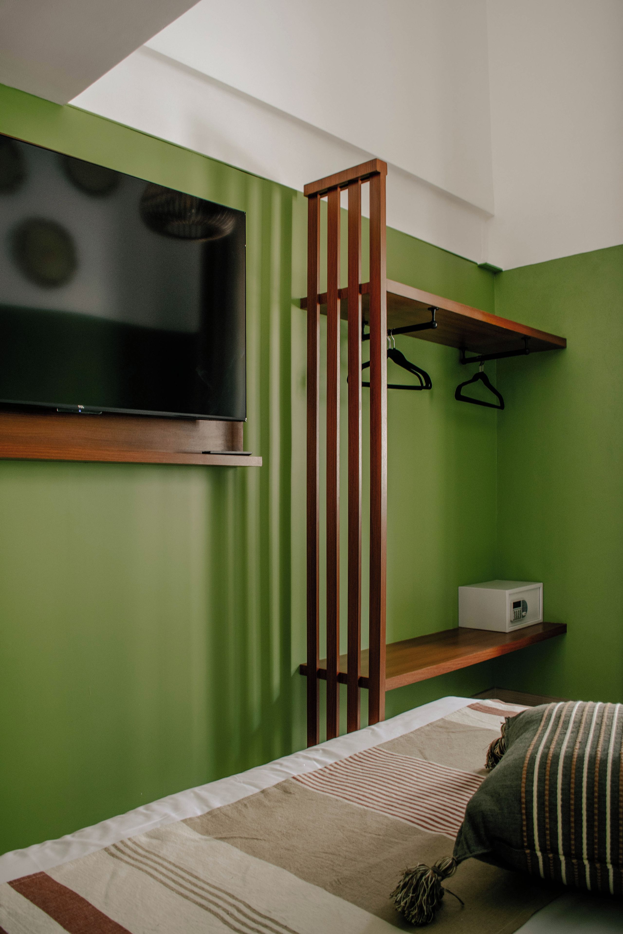 Una habitación compacta con una cama, un televisor montado en la pared, un estante de madera y una pared verde.